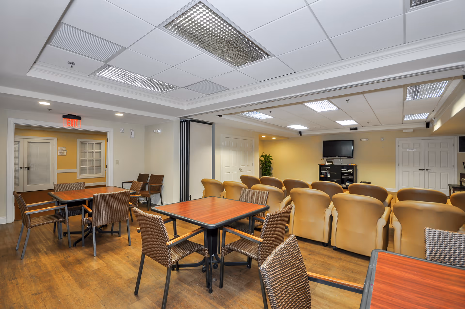 A senior living community common room with tables, wicker chairs and rows of tan recliners facing a wall-mounted TV.
