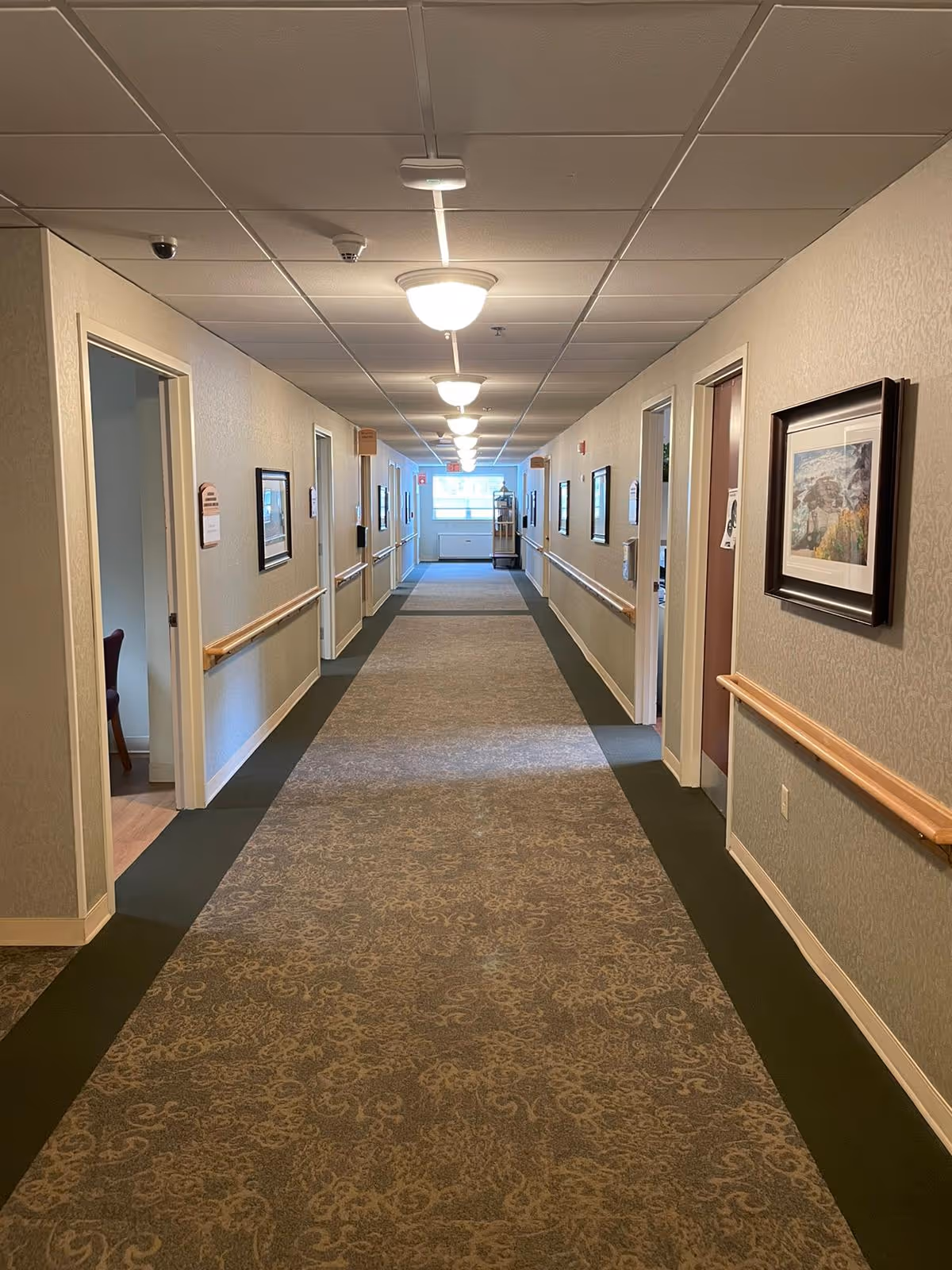 A long, well-lit hallway in a nursing and rehabilitation center with patterned carpet flooring and handrails on both sides. Several doorways and framed pictures are visible along the walls, with ceiling lights evenly spaced overhead. A window at the far end lets in natural light.