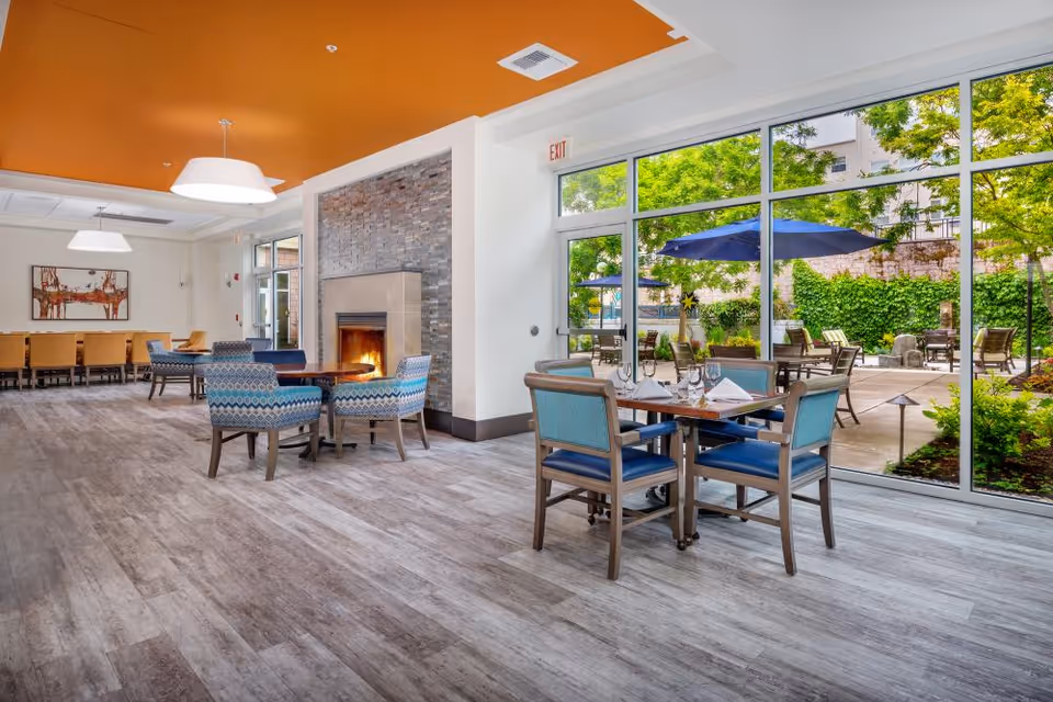 Bright communal dining and lounge area with tables and chairs, a fireplace, and large windows overlooking an outdoor patio.