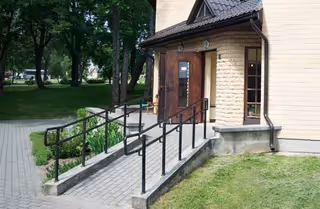 Brick building entrance with a sloped ramp and metal railing, surrounded by green landscaping.