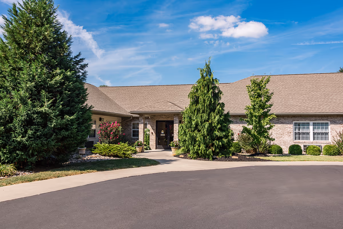 Front entrance of a single-story brick senior living building with landscaped shrubs and trees under a blue sky.