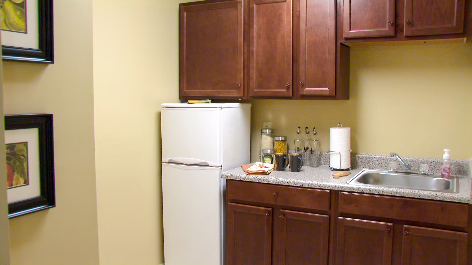 A small kitchen area with wooden cabinets, a white refrigerator, a countertop with a sink, paper towel holder, soap dispenser, two mugs, glass containers with dry food, and framed pictures on the adjacent wall.