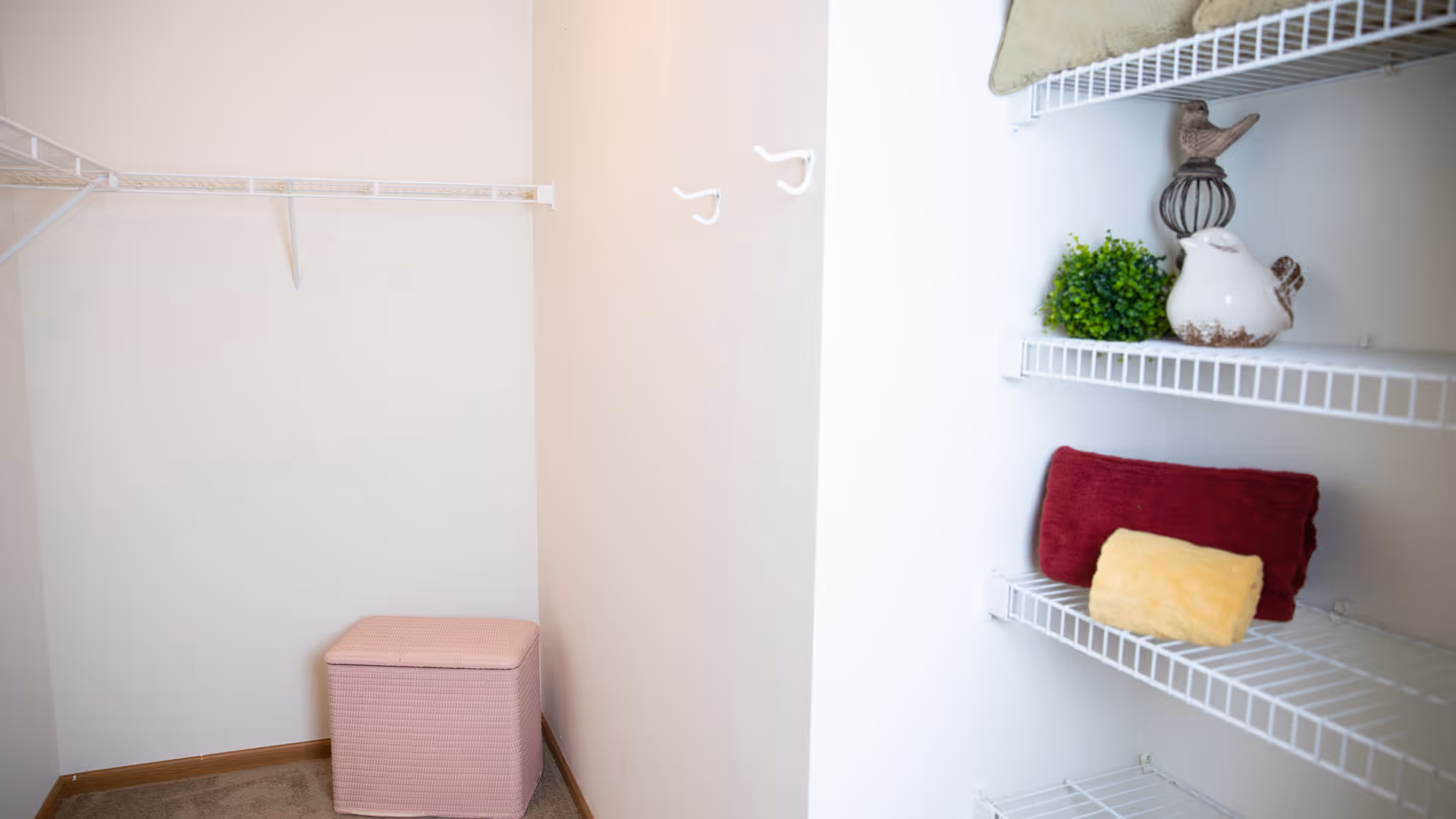 A small walk-in closet with white wire shelving on the walls. There are two hooks on the wall and a pink storage ottoman on the floor. On the shelves, there are decorative items including a small green plant, two ceramic bird figurines, and folded towels in red and yellow.