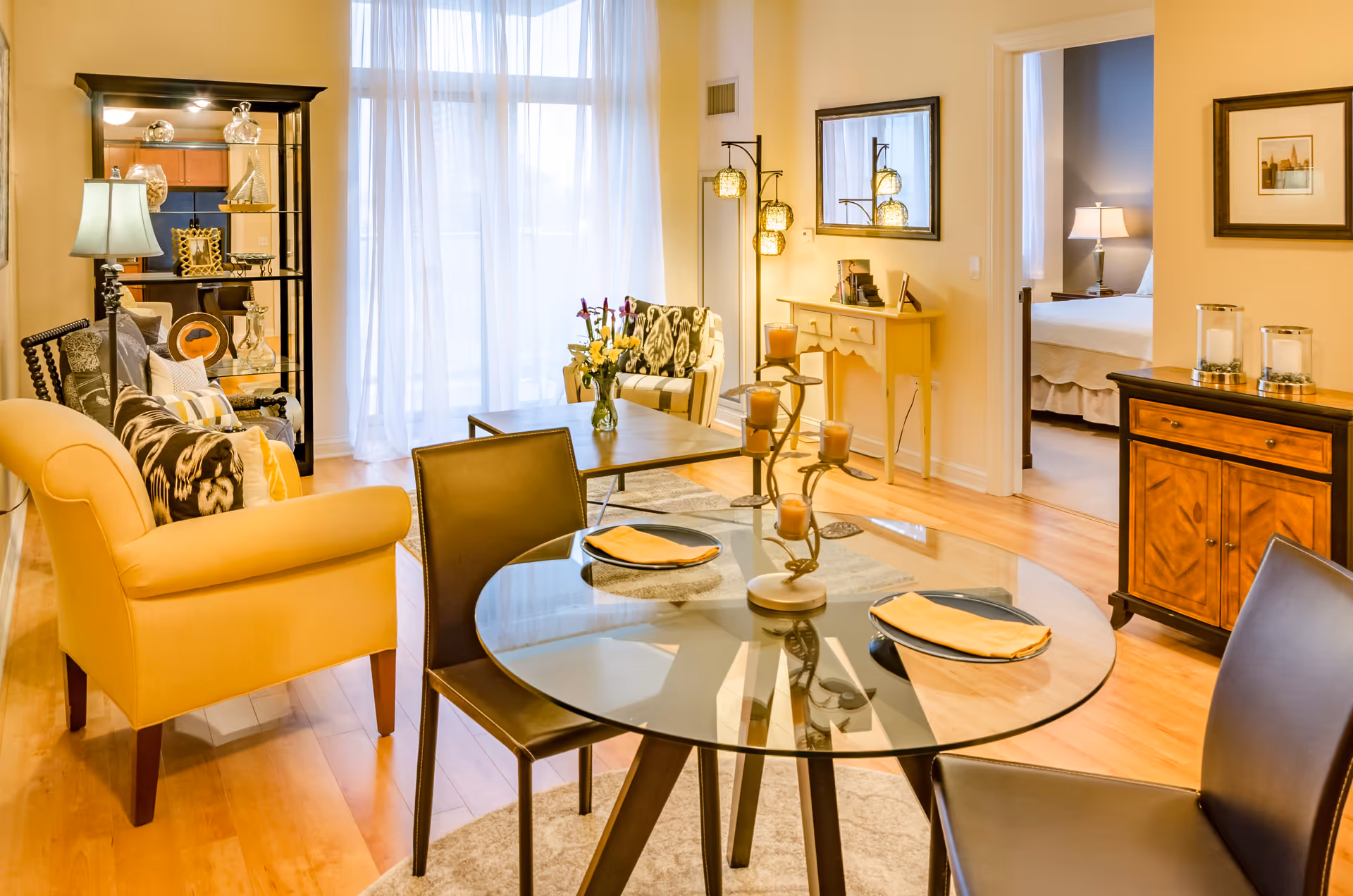 A bright and cozy living and dining area with a glass round dining table set with two place settings, a yellow armchair, a patterned sofa, a coffee table with a vase of flowers, a tall black shelving unit with decorative items, and a doorway leading to a bedroom with a bed and a lamp. The room has wooden floors, light-colored walls, and sheer white curtains covering large windows.