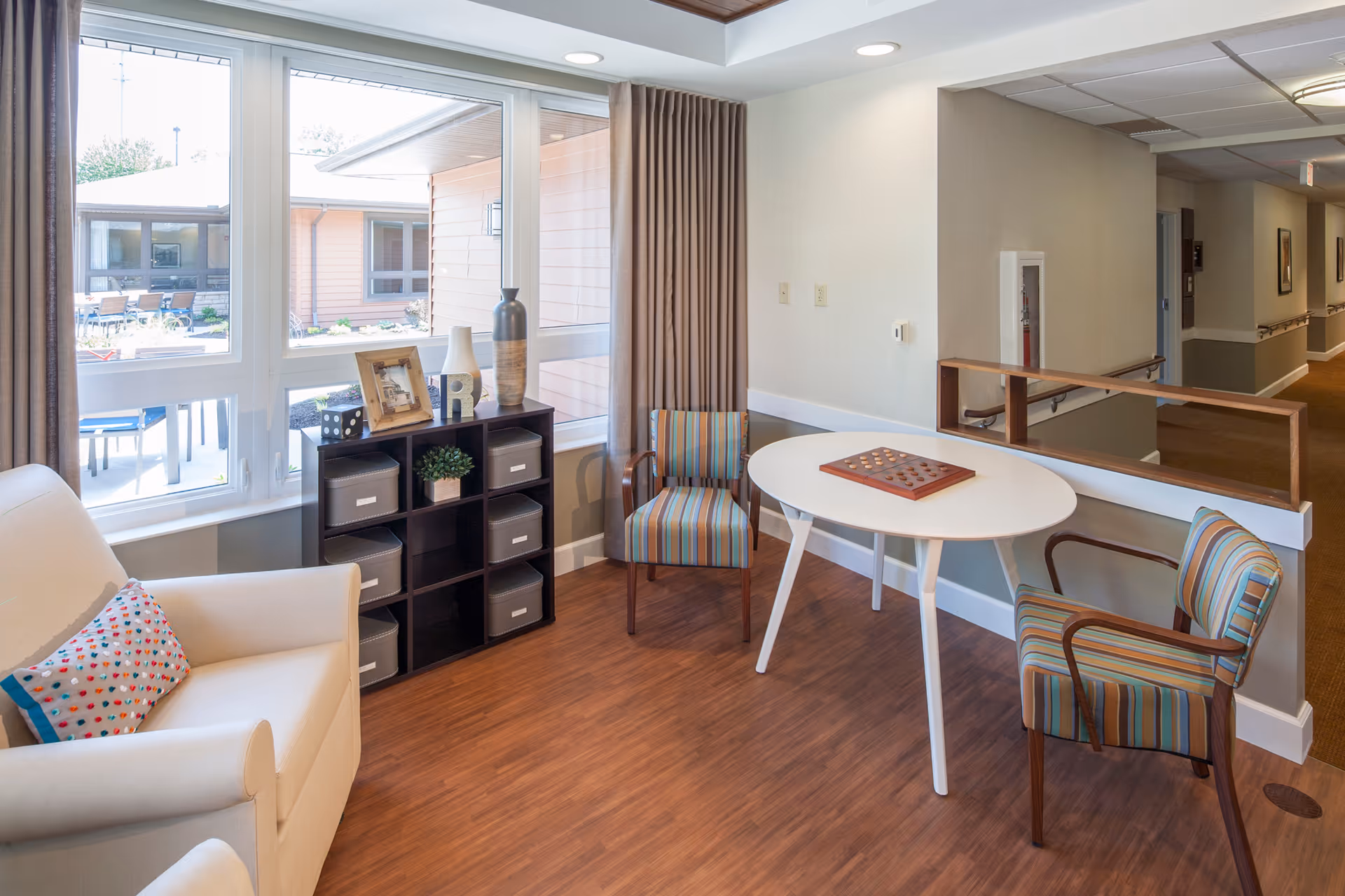 A cozy seating area in a senior living facility with a white armchair featuring a colorful heart-patterned pillow, two striped armchairs, and a white oval table with a checkers game on it. There is a black cubby shelf with decorative items and storage bins next to large windows with beige curtains, allowing natural light to brighten the room. The space has wood flooring and a hallway visible in the background.