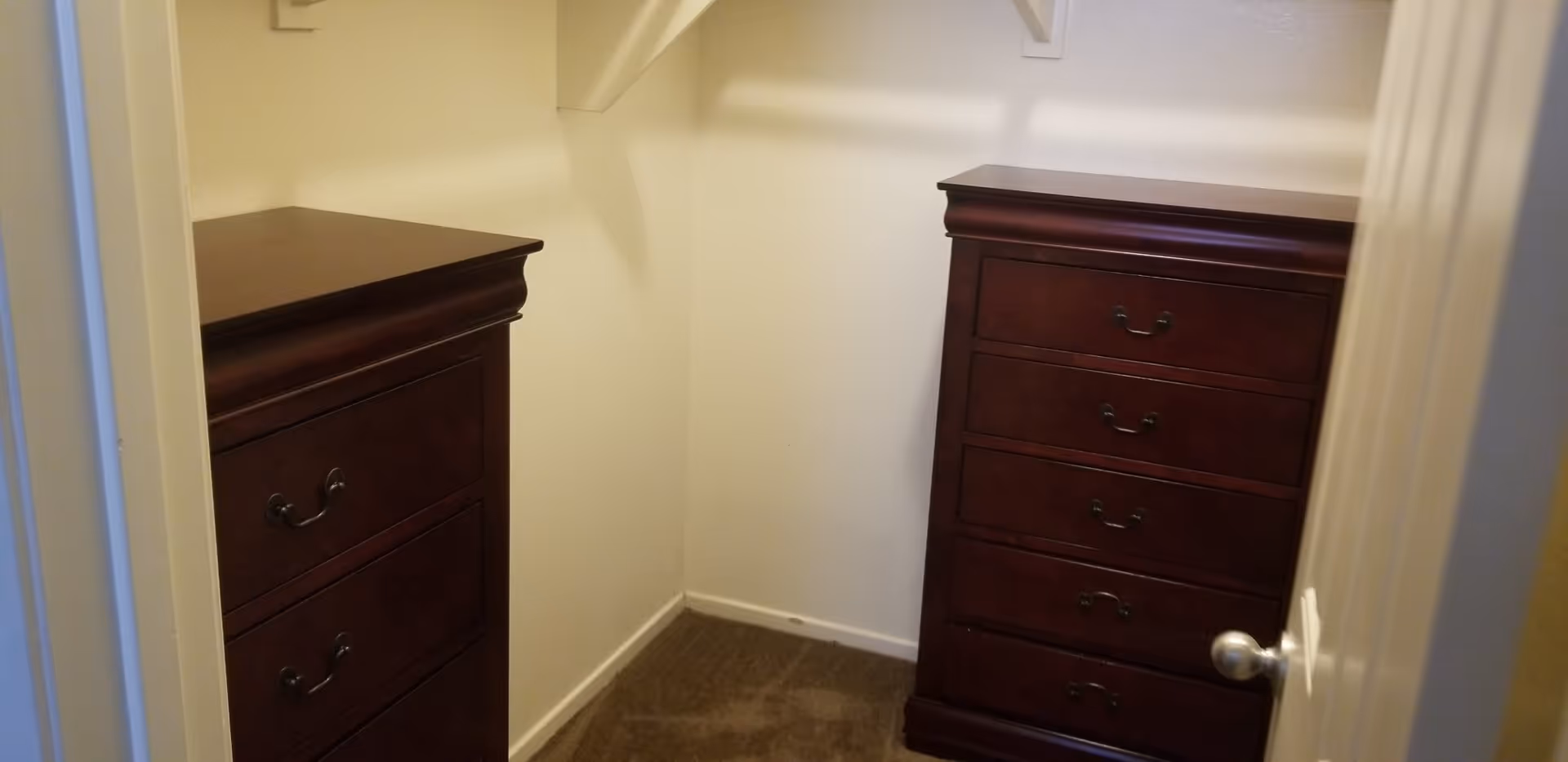 A small walk-in closet with beige walls and carpeted floor containing two dark wooden dressers with multiple drawers, one on each side of the closet.
