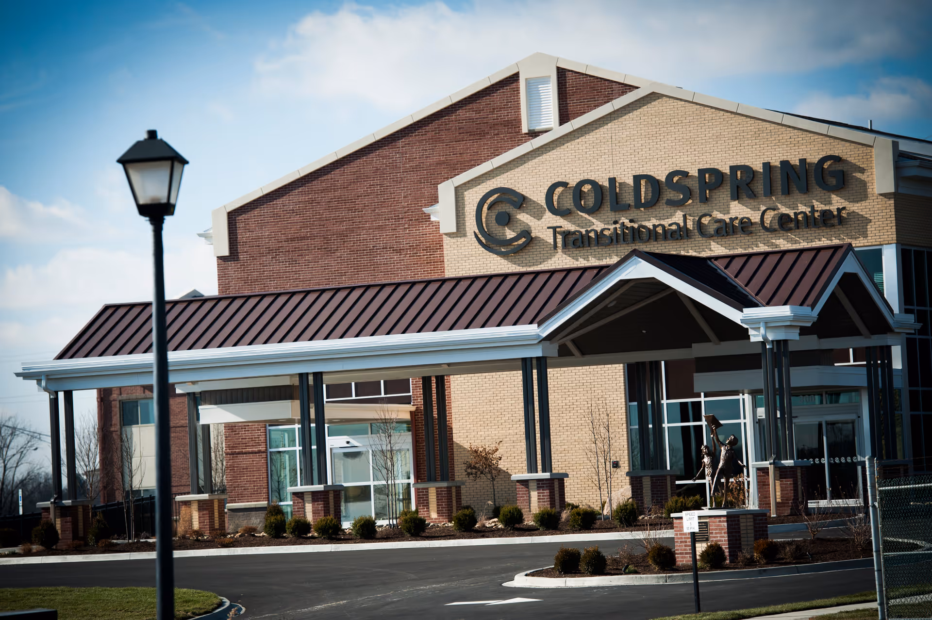 Exterior view of the Coldspring Transitional Care Center building with a covered entrance, a street lamp in the foreground, and a clear sky.