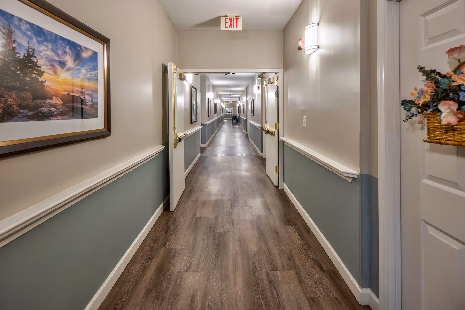 A long, well-lit hallway in a senior living facility with wood flooring and light-colored walls painted beige on top and blue on the bottom. The hallway features several doors on both sides, some open, and framed landscape artwork on the walls. A basket with artificial flowers is hanging on a door to the right, and an exit sign is visible at the far end of the corridor.