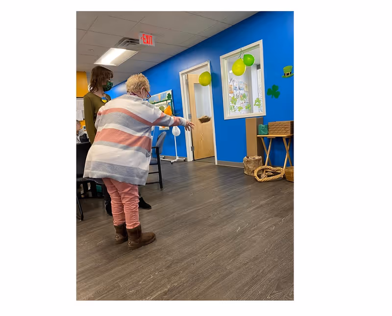 An elderly woman wearing a striped sweater and pink pants is throwing a bean bag in an indoor room with blue walls decorated with green and yellow balloons and shamrock cutouts. Another person wearing a mask stands nearby watching. The room has wood flooring and doors with windows.