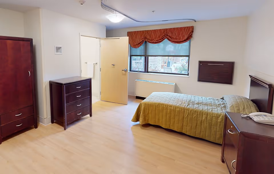 A senior living bedroom with a single bed, nightstand, dresser and armoire, a window with a valance, and an open door to an adjoining bathroom.