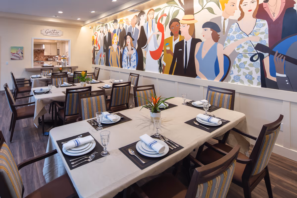 Bright dining room with multiple set tables and chairs and a large colorful mural on the wall.