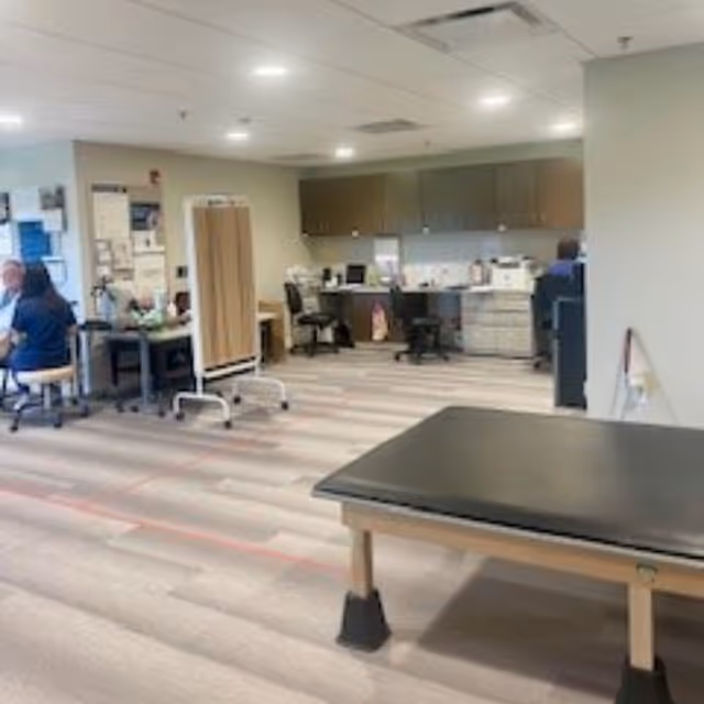 Open interior therapy room with a padded treatment table in the foreground and desks with staff workstations along the back wall.