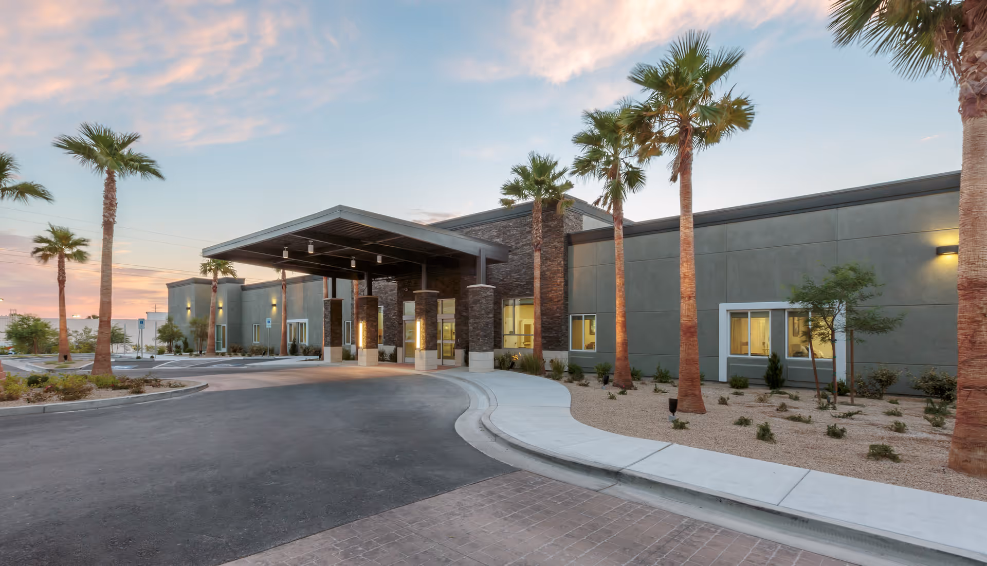 Exterior view of a modern single-story building with a covered entrance, surrounded by palm trees and desert landscaping under a partly cloudy sky at sunset.