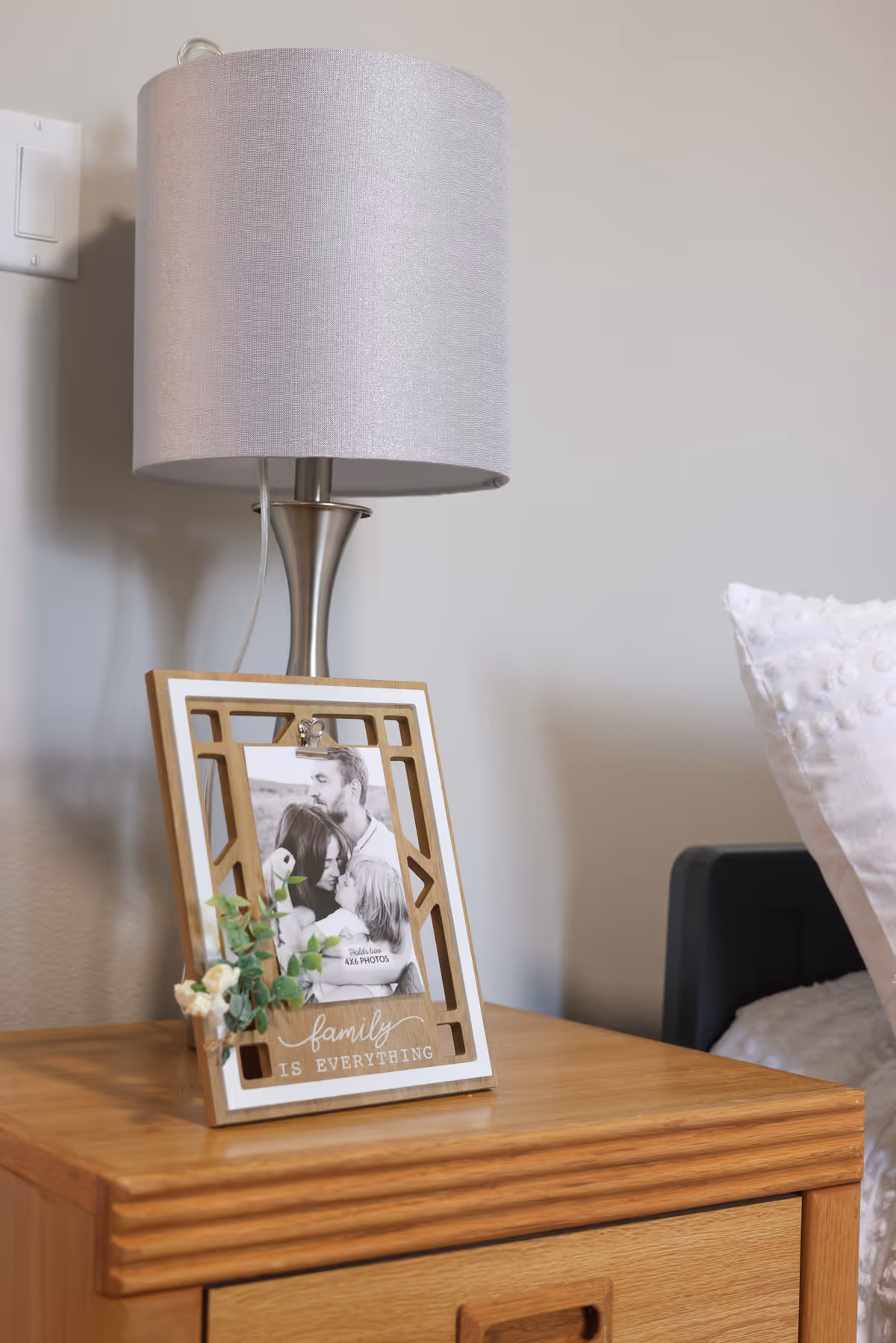 Wooden nightstand holding a table lamp and a framed family photo next to a bed pillow.