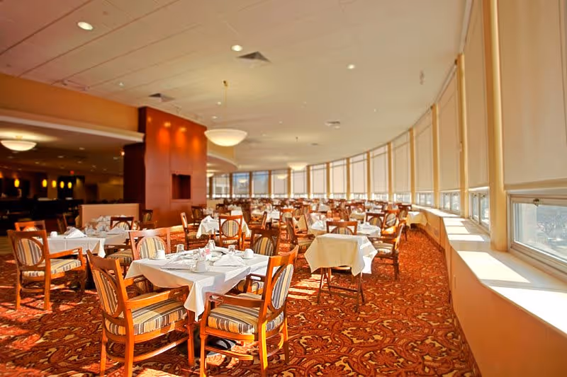 A spacious dining room with multiple tables covered in white tablecloths, each set with plates, cups, and silverware. The room features large windows with blinds partially drawn, allowing natural light to fill the space. The carpet has a red and orange patterned design, and wooden chairs with striped cushions surround the tables.