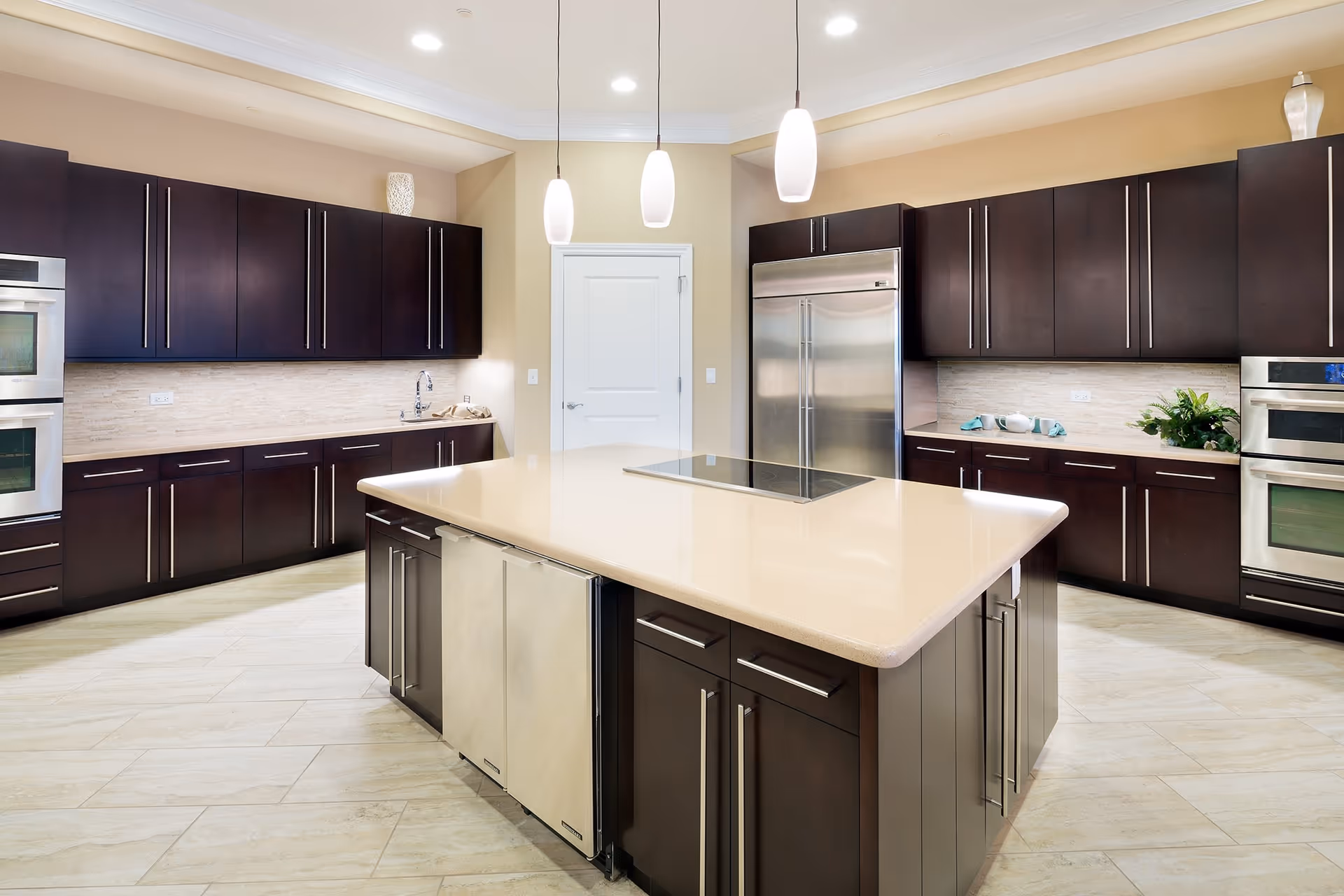A modern kitchen with dark wood cabinets, stainless steel appliances including a refrigerator and double ovens, a large beige island countertop with built-in stovetop, three pendant lights hanging from the ceiling, and light-colored tile flooring.