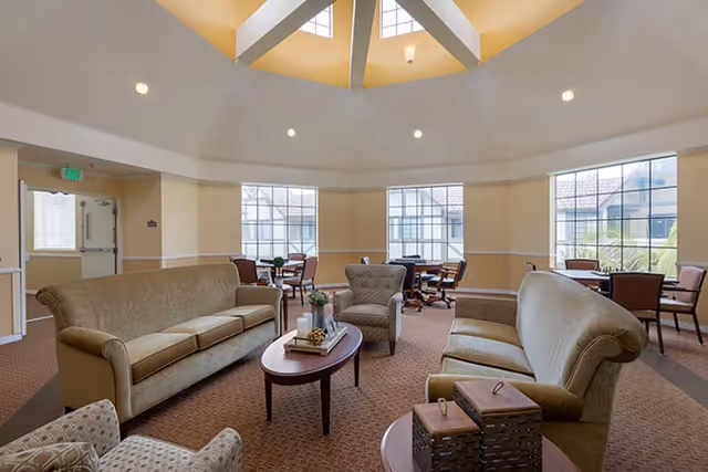 A spacious senior living common area with beige sofas and armchairs arranged around a wooden coffee table. The room has large windows letting in natural light and a high ceiling with a skylight. Several small tables and chairs are placed near the windows.
