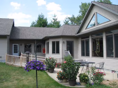 A single-story senior living building with a front porch, railing, and potted flowers along the walkway.