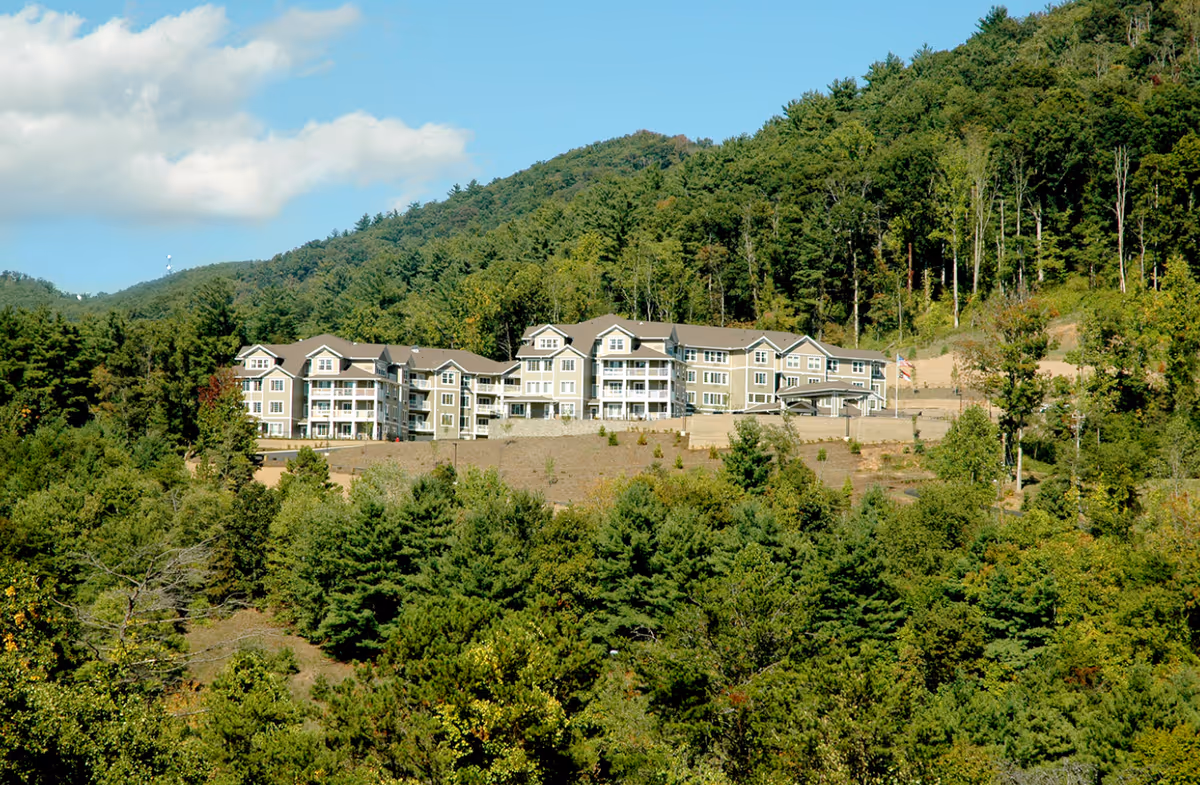 A large multi-story residential building situated on a hillside surrounded by dense green trees and forested mountains under a partly cloudy blue sky.
