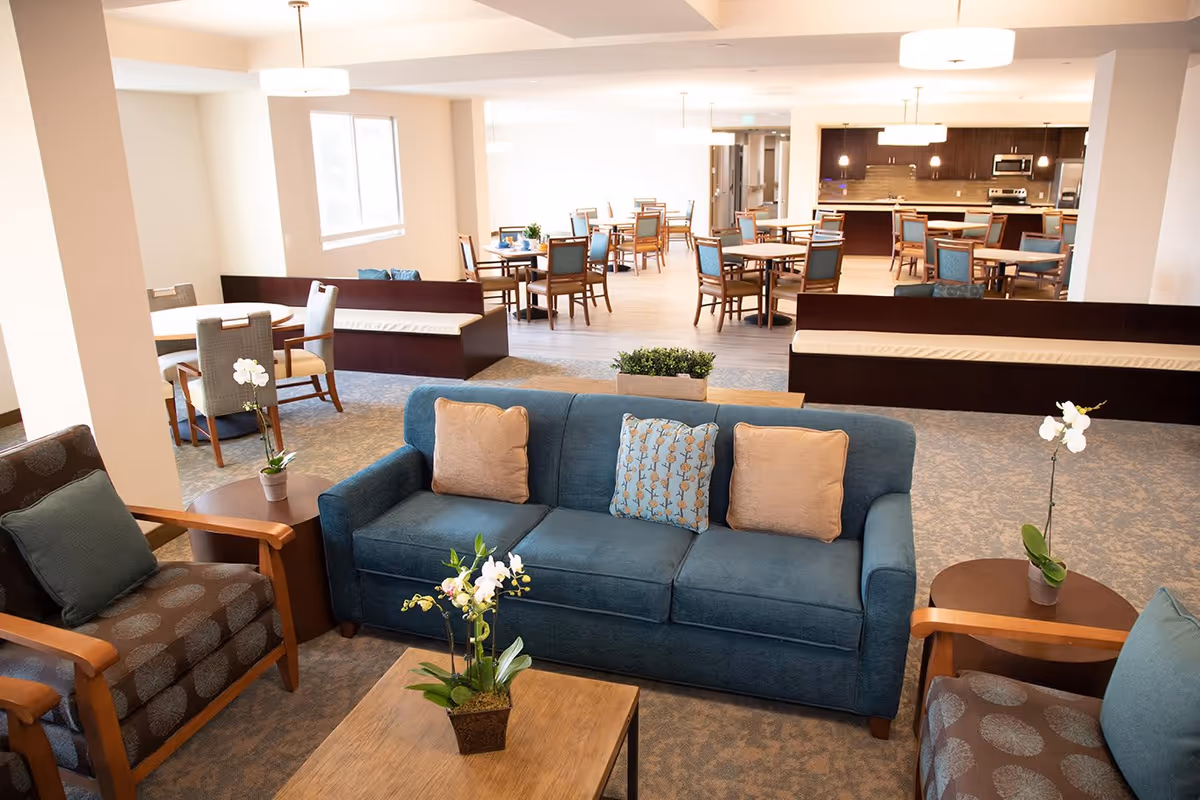 A bright and spacious senior living common area featuring a blue sofa with three cushions, two patterned armchairs with blue pillows, wooden side tables with white orchid plants, and multiple dining tables and chairs in the background near a kitchen area with dark cabinetry and stainless steel appliances.