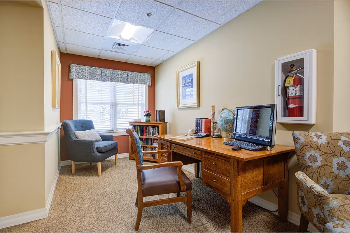 Small cozy sitting and work area with a wooden desk and computer, upholstered chairs, bookshelf, and a window with a valance.