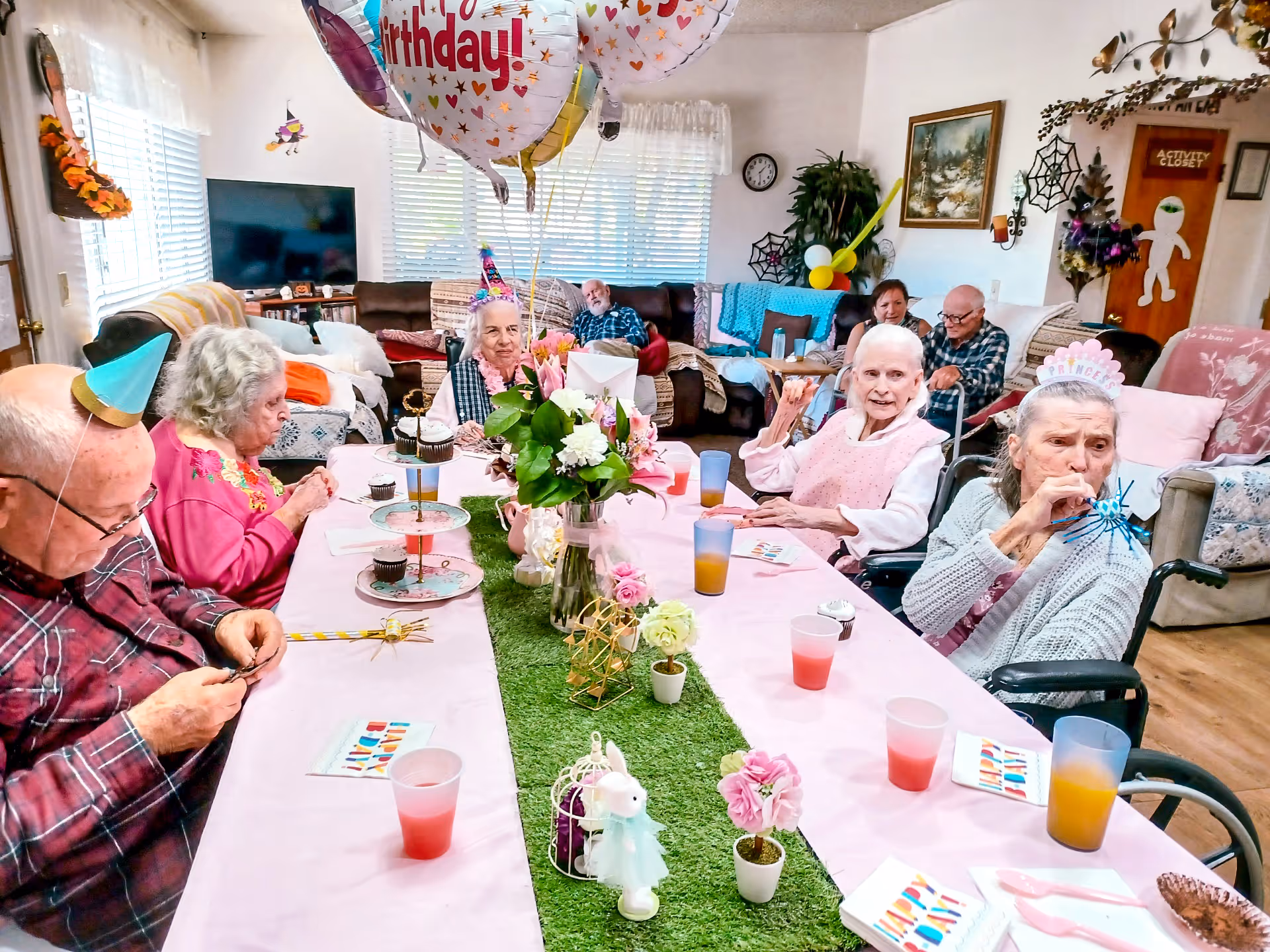 A group of elderly people sitting around a long table decorated with a pink tablecloth, green grass-like runner, flowers, and birthday decorations. Some are wearing party hats and birthday accessories, with balloons overhead. The room has couches, a TV, and festive decorations on the walls, indicating a birthday celebration in a communal living area.