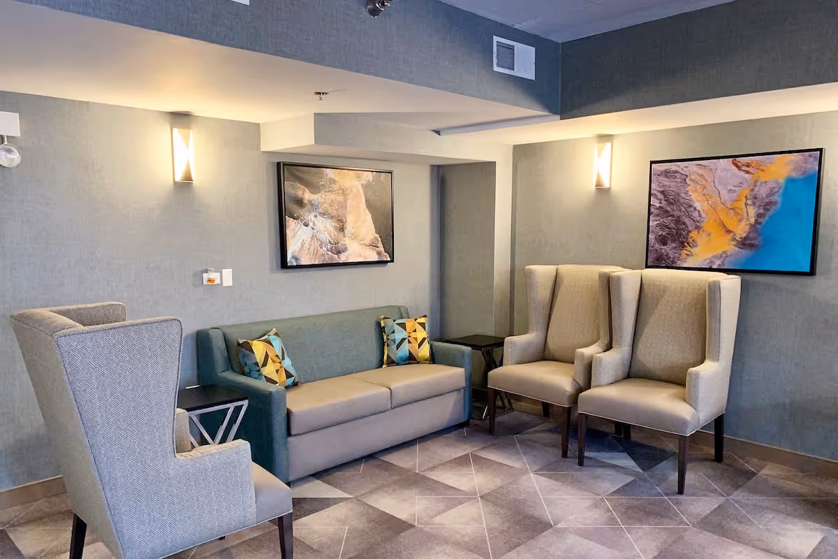 A cozy seating area in a supportive living facility featuring a gray sofa with two patterned cushions, two beige wingback chairs, and a gray armchair. The walls are painted light gray and adorned with two framed abstract art pieces. The floor has a geometric tile pattern, and soft wall-mounted lights illuminate the space.