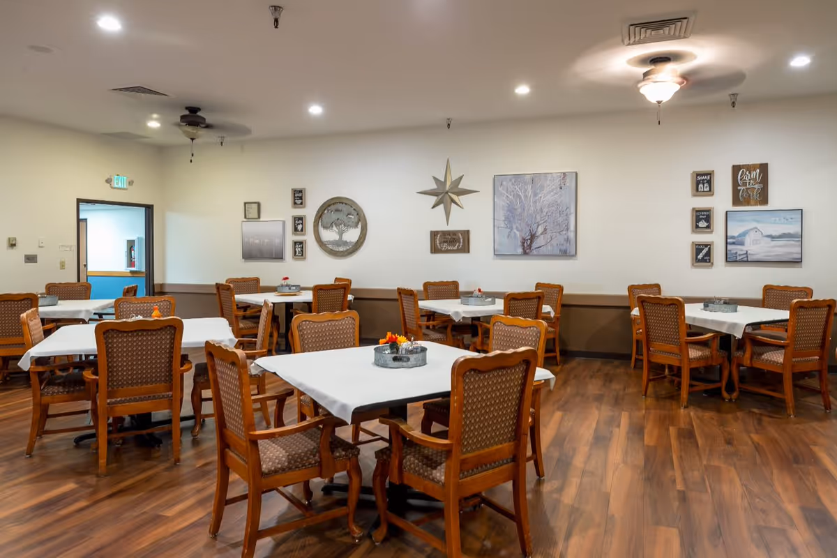 A dining room with several wooden tables covered with white tablecloths and surrounded by wooden chairs with patterned cushions. The room has wooden flooring, white walls decorated with various framed pictures and wall art, and ceiling fans with lights. There is an exit door visible on the left side.