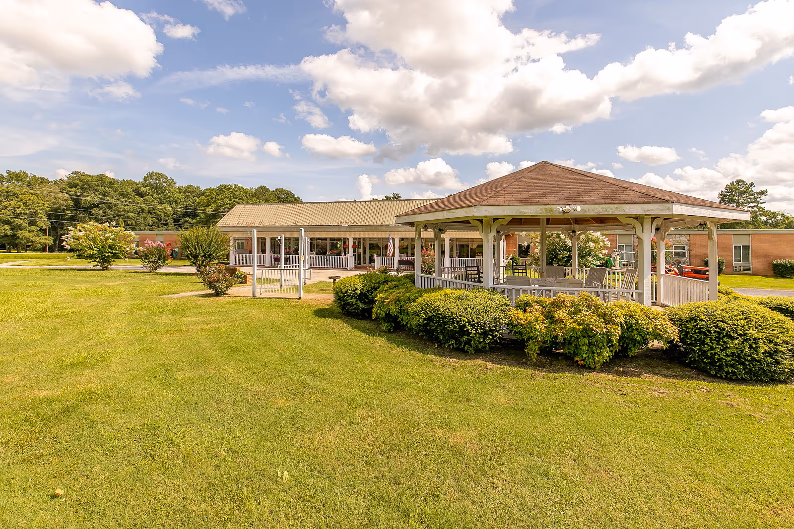 Outdoor view of a senior living facility with a large green lawn, a white gazebo surrounded by bushes, and a single-story brick building in the background under a partly cloudy sky.