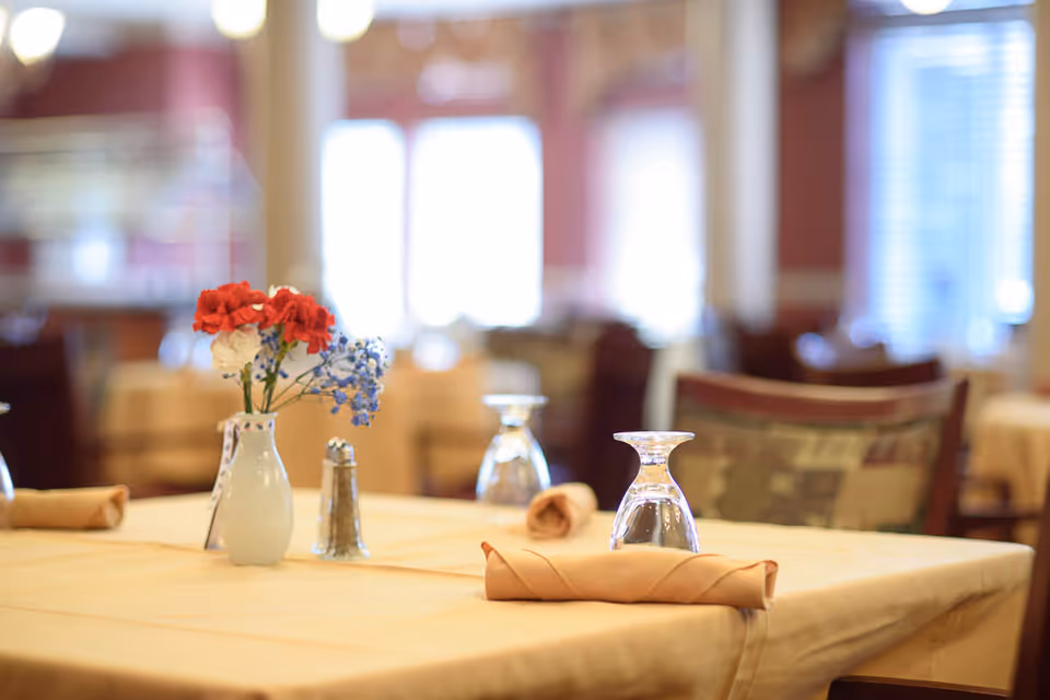 A dining table set with a beige tablecloth, a small vase with red, white, and blue flowers, a salt shaker, upside-down drinking glasses, and rolled beige napkins in a softly lit dining room with blurred chairs and windows in the background.