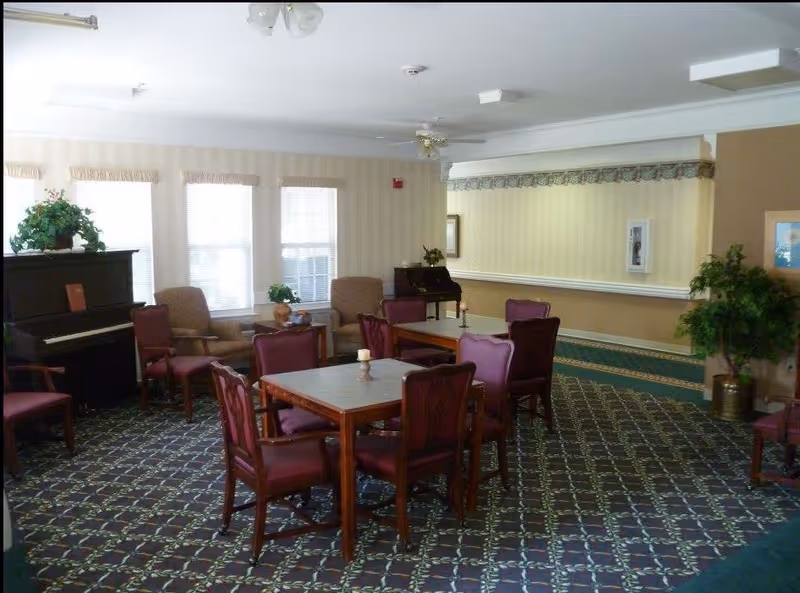 A senior living facility common area with multiple tables and chairs arranged on a patterned carpet. There are armchairs near the windows, a piano with a plant on top, and framed artwork on the walls. The room has soft lighting and a cozy atmosphere.