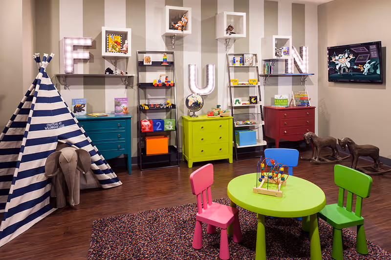 A colorful children's playroom with a small round green table and three small chairs in pink, blue, and green. There is a striped blue and white teepee with a stuffed elephant toy next to it. The wall has shelves with toys, books, and decorative letters spelling 'FUN' illuminated with lights. A TV is mounted on the wall showing an animated cartoon, and two dog sculptures are on the floor near the TV.