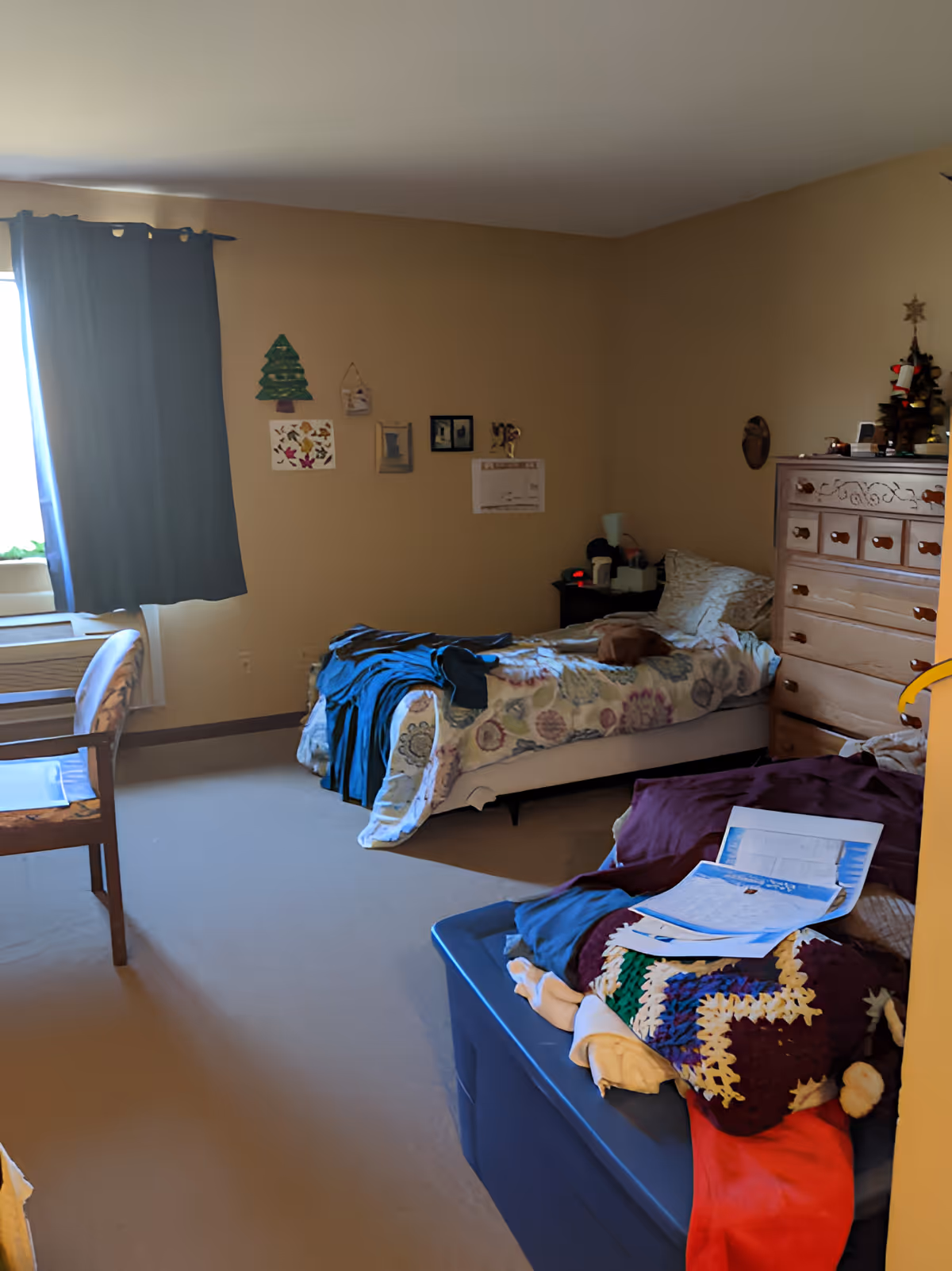 A small bedroom with a single bed covered with a floral blanket and some clothes on it. There is a wooden dresser with a small Christmas tree decoration on top. A blue storage bin with a crocheted blanket and papers on it is in the foreground. A wooden chair is near a window with blue curtains. The walls have a few small decorations and pictures.