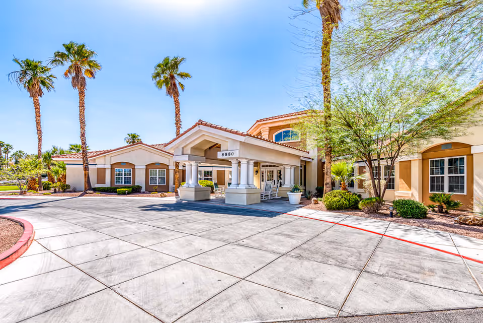 Exterior view of Spring Valley Memory Care facility with a covered entrance supported by columns, surrounded by palm trees and desert landscaping under a clear blue sky.