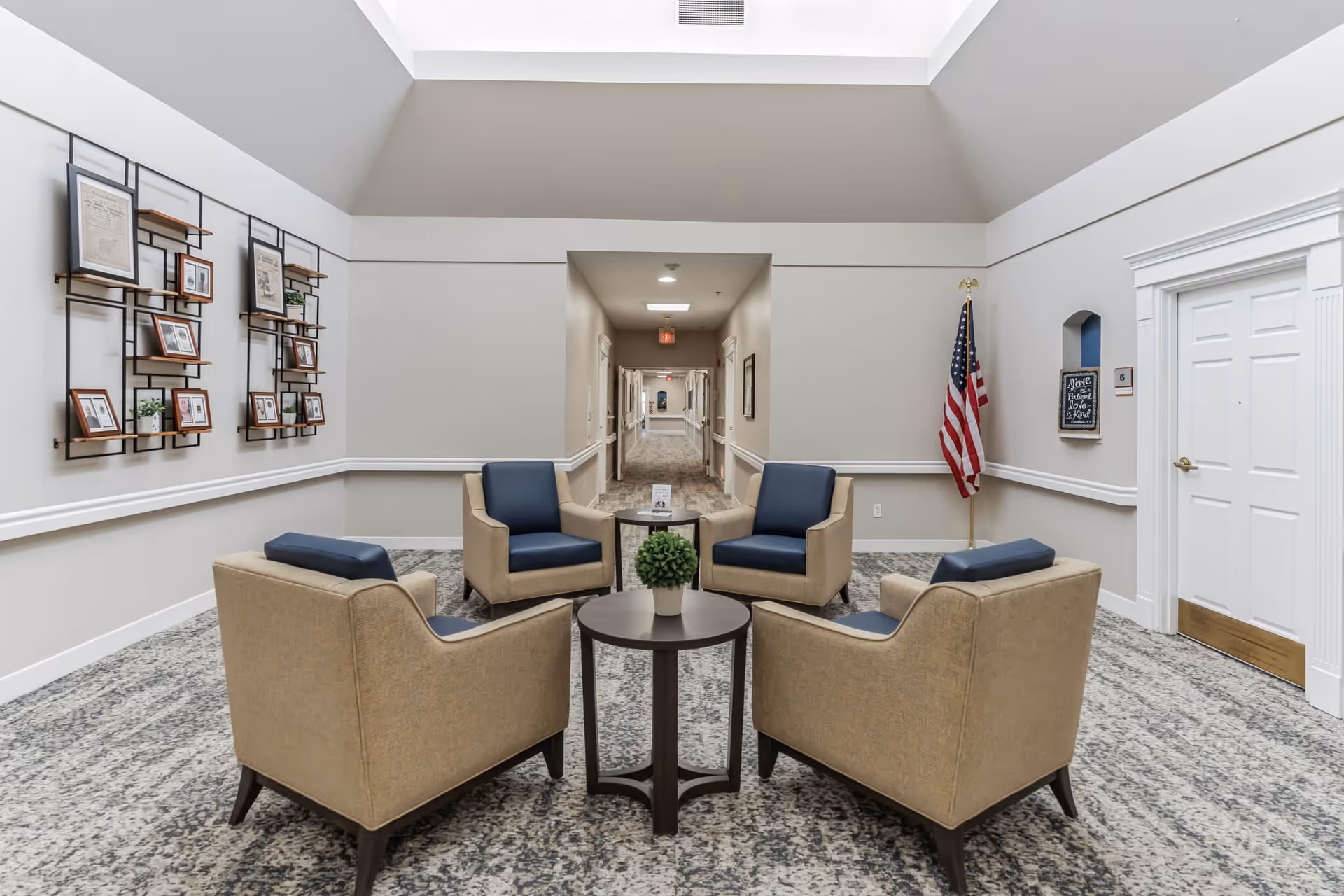 A seating area in a senior living facility with four beige armchairs featuring dark blue cushions arranged around two small round tables with a potted plant on one. The walls are light gray with white trim, decorated with framed pictures and an American flag in the corner. A hallway extends in the background with doors on either side and a carpeted floor.