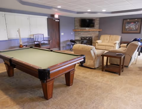 Recreation lounge with a pool table in the foreground, a seating area of recliners and a sofa near a fireplace and wall-mounted TV.