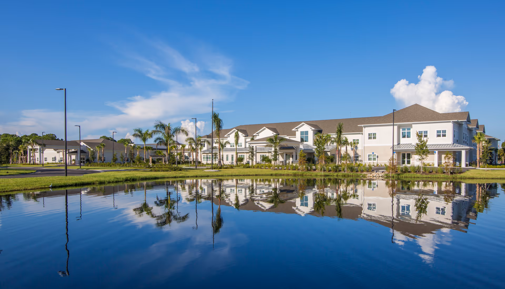 A large, modern senior living facility building with white and beige exterior walls and a gray roof, surrounded by palm trees and landscaping, reflected in a calm pond in the foreground under a clear blue sky.