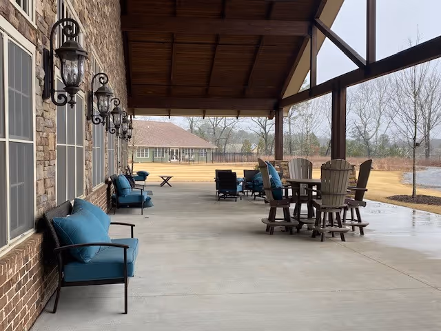 Covered outdoor patio with cushioned chairs and tables along a brick wall overlooking a lawn and pond.