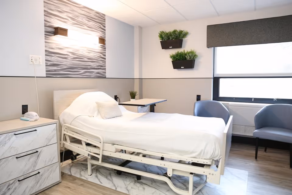 A clean and modern nursing facility room with a hospital bed covered in white linens, a small pillow, a bedside table with a telephone, two gray chairs near a large window with a gray valance, and two small wall-mounted planters with green plants.