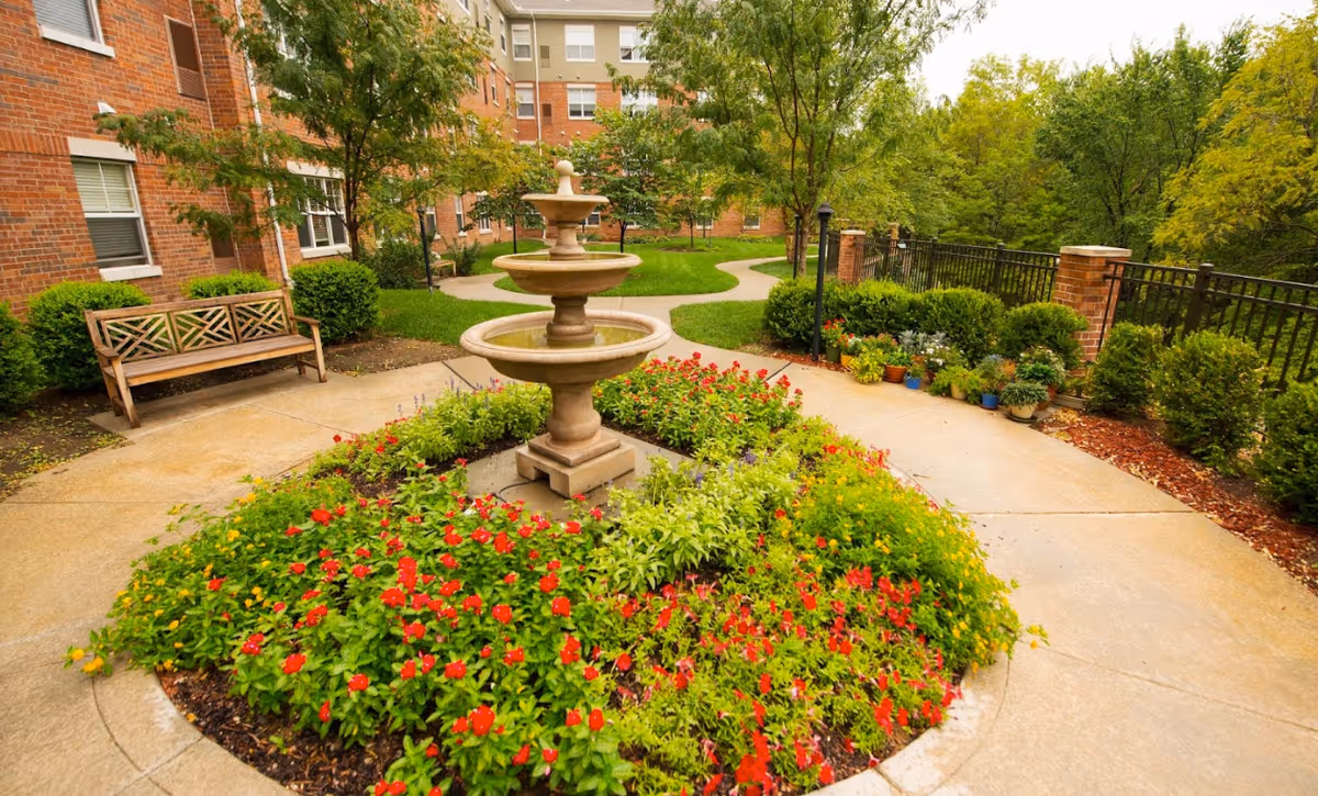 A landscaped outdoor garden area at The Gardens at Creekside featuring a three-tiered stone fountain surrounded by vibrant red and yellow flowers. There is a wooden bench on the left side, a paved walkway winding through green grass and trees, and a black metal fence with brick pillars on the right side.