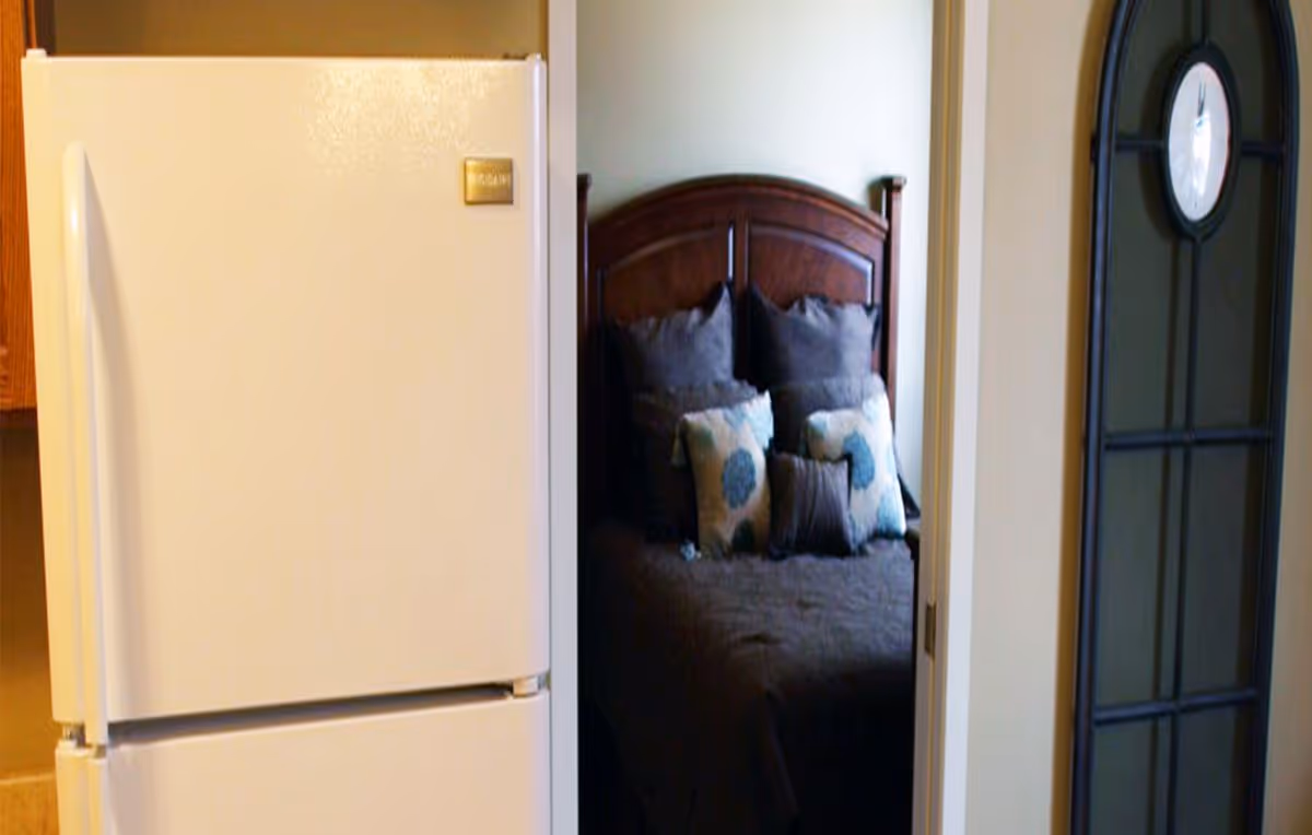 View of a white refrigerator in a kitchen area with a doorway leading to a bedroom. The bedroom has a wooden headboard and a bed with dark bedding and decorative pillows. To the right of the doorway is a tall wall clock with a black frame.