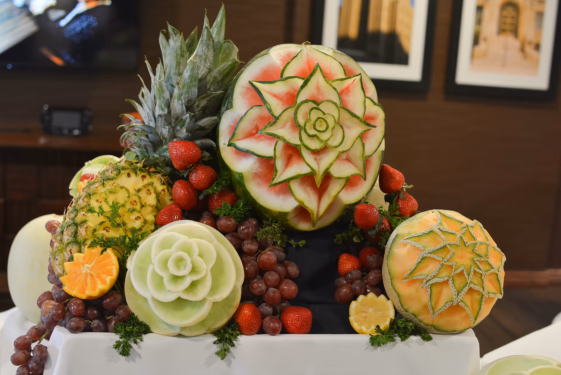 A decorative arrangement of carved fruits including watermelon, cantaloupe, honeydew melon, pineapple, strawberries, grapes, and an orange slice, displayed on a white tablecloth with framed pictures on a brown wall in the background.