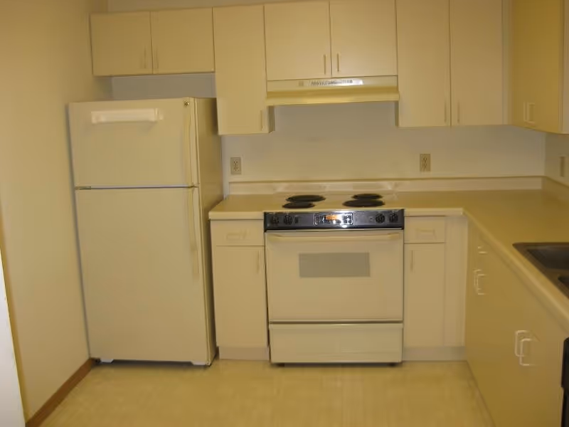 A small kitchen with white cabinets, a white refrigerator, an electric stove with four burners, and a range hood. The countertops are light-colored, and the floor has a light-colored tile pattern.