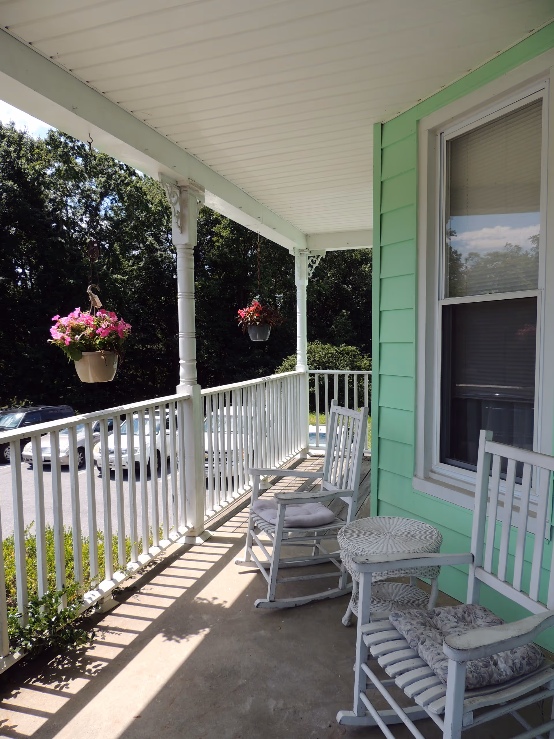A covered porch with white wooden railing and two white rocking chairs with cushions. There is a small round white wicker table between the chairs. Two hanging flower pots with pink flowers are suspended from the porch ceiling. The exterior wall of the building is painted light green, and a window with closed blinds is visible. In the background, there are trees and parked cars.