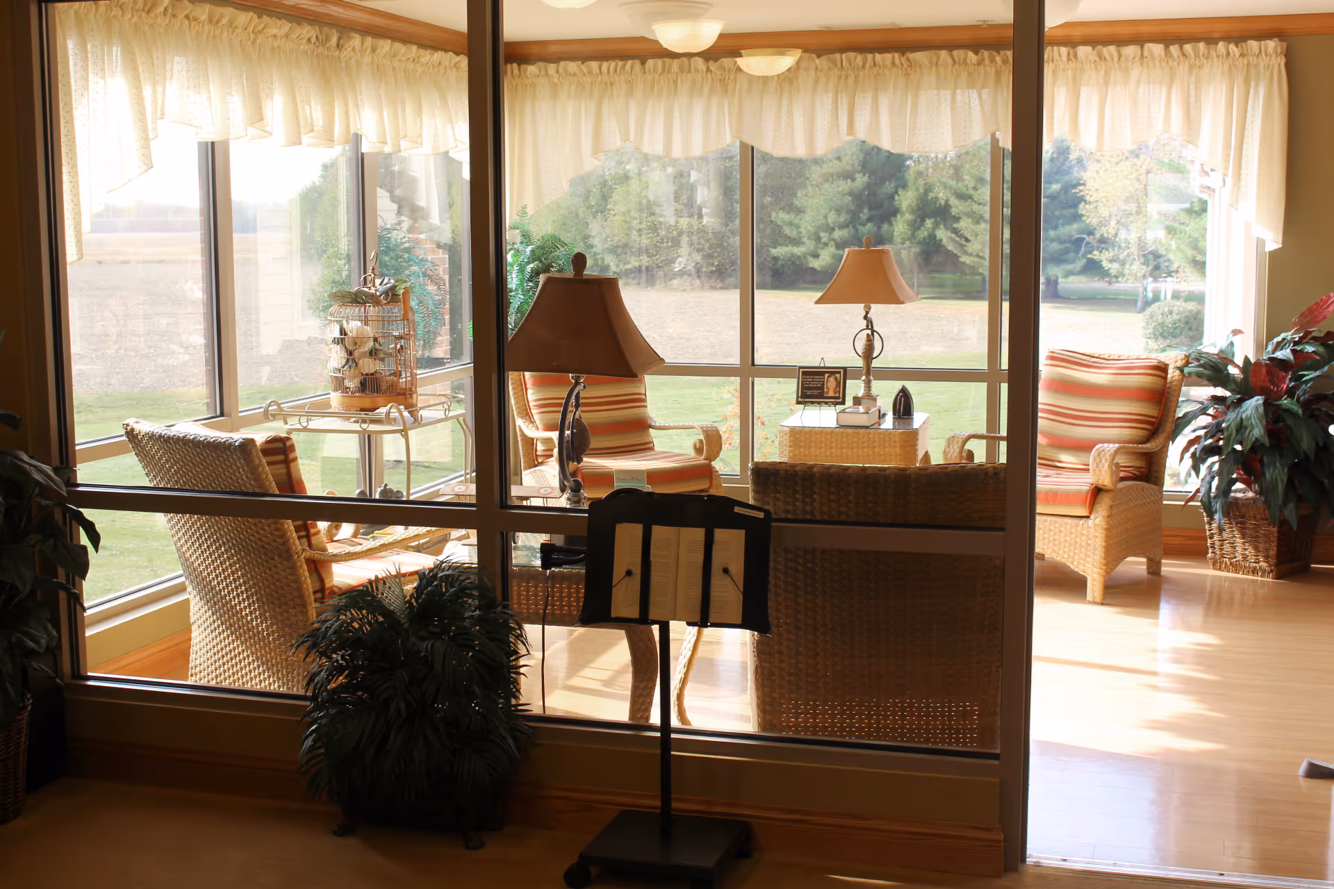 A cozy sunroom with large windows overlooking a grassy outdoor area. The room contains wicker chairs with striped cushions, a small table with a lamp, and decorative plants. Light curtains frame the windows, and a music stand is visible in the foreground.