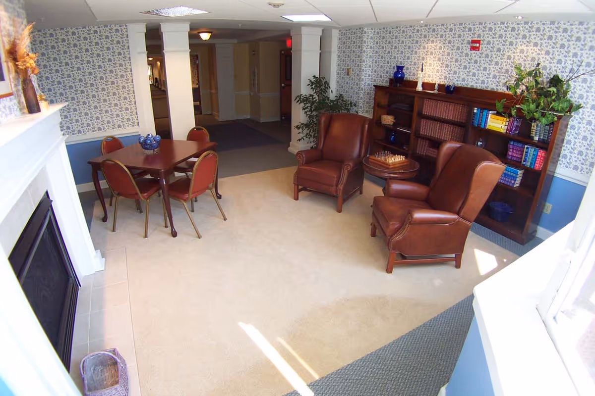 Carpeted common room with two leather armchairs around a small table, a bookshelf, a dining table with four chairs, and a fireplace.