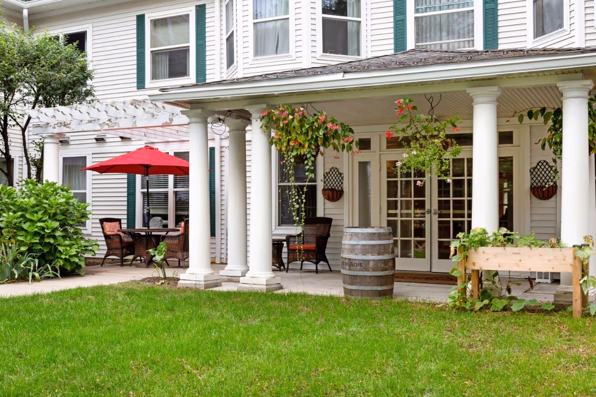Outdoor patio area of a senior living facility with white columns supporting a covered porch. There are hanging flower baskets, a wooden barrel, a raised garden bed, and a seating area with a table and chairs under a red umbrella. The building has white siding and green shutters on the windows.
