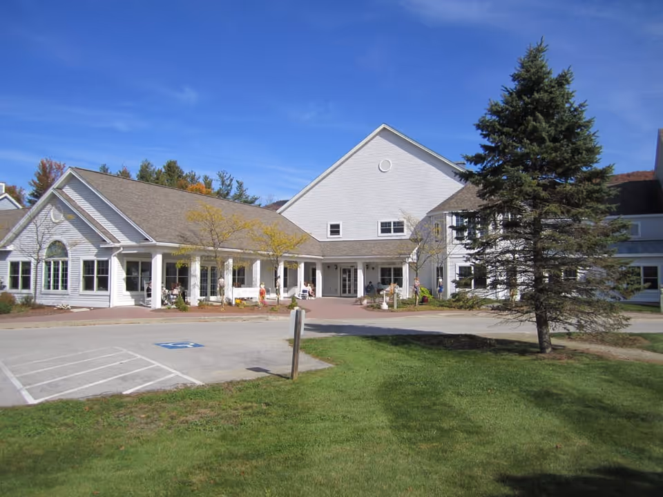 Front exterior of a white senior living facility with a covered entrance, parking area, and a pine tree on the lawn.