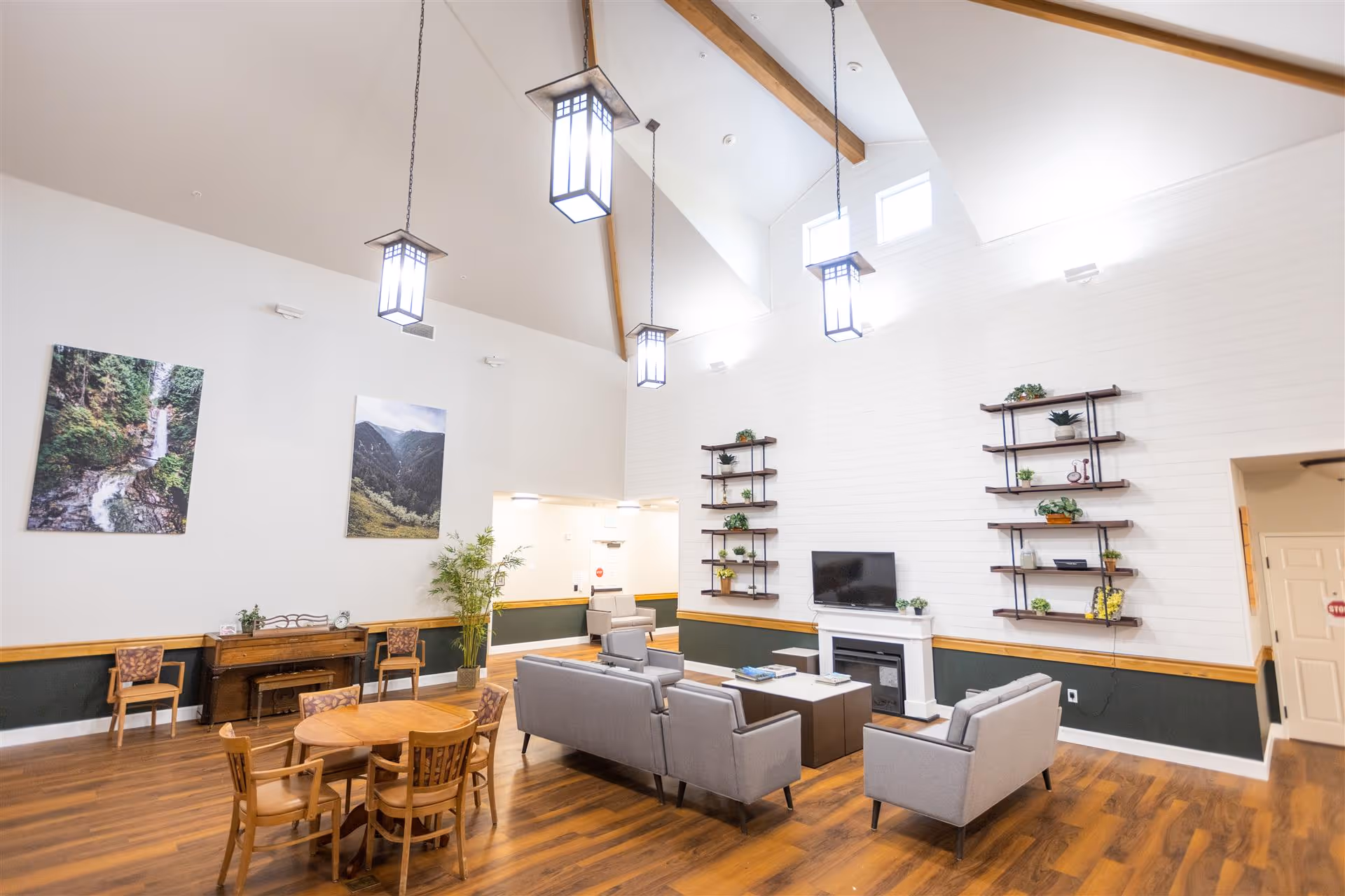Spacious senior living common room with a vaulted ceiling, hanging pendant lights, a seating area with sofas around a TV and fireplace, and a round dining table.