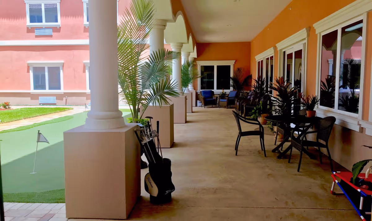 Covered outdoor patio area with columns, potted plants, and several chairs and tables along the wall. A small putting green with a flag is visible on the left side, and the exterior of a pink building with windows is in the background.